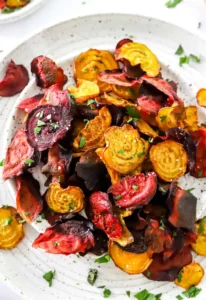 Crispy baked beet chips on a white plate with green herbs on top with another plate of beet chips behind it.
