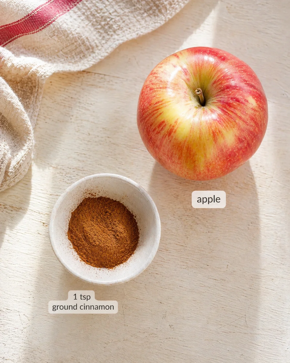 red-yellow apple next to a small bowl of ground cinnamon on a light wooden table with a woven cloth