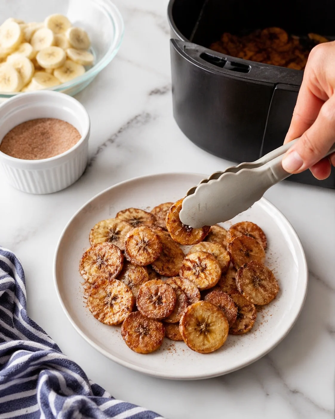 Golden-brown banana chips on a white plate, tongs lifting a slice, background shows sliced bananas and brown sugar.