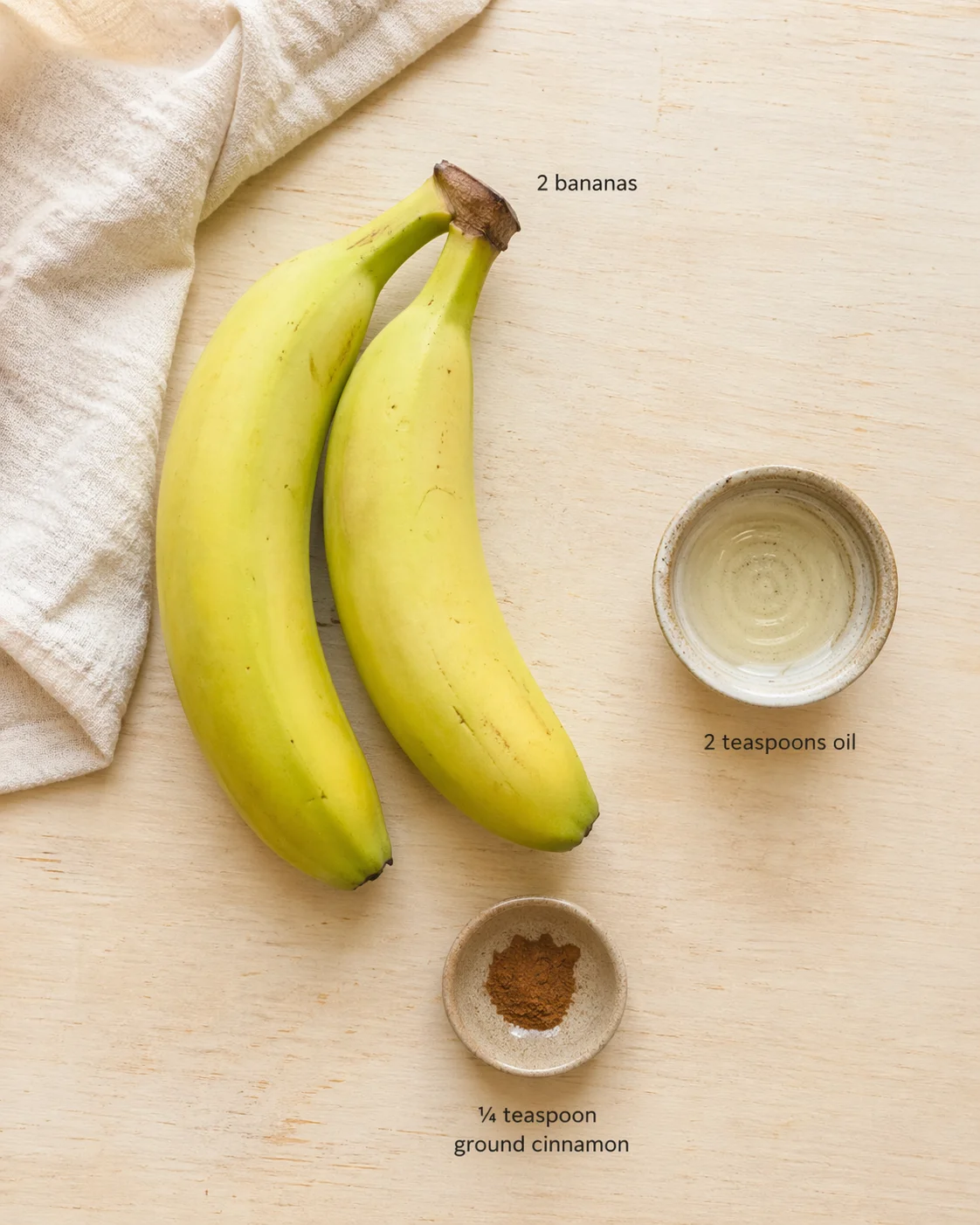 Two bananas on a light wooden surface with labeled bowls of oil and ground cinnamon.