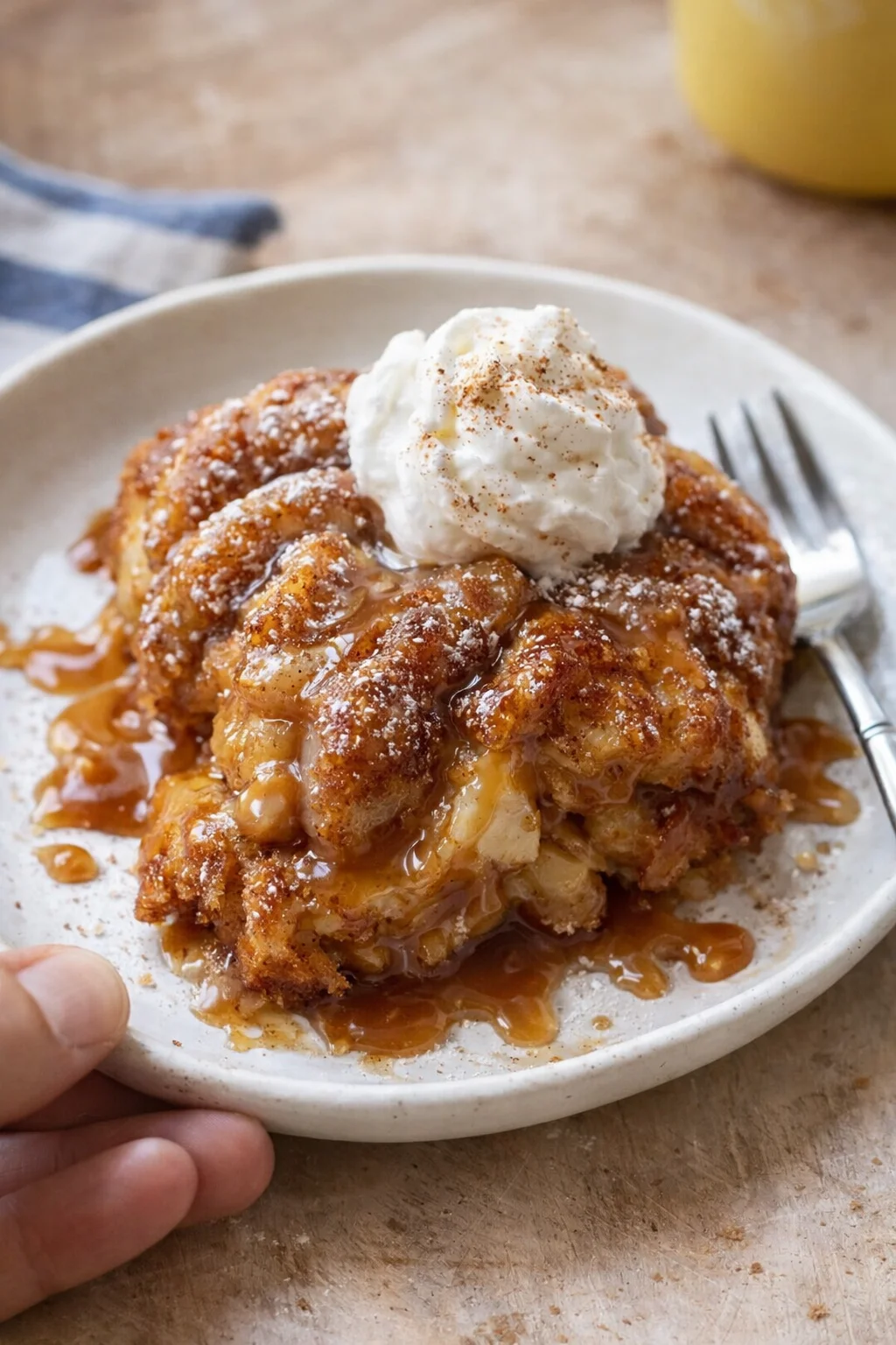 Warm caramel-drizzled monkey bread on a white plate, topped with whipped cream and cinnamon dust.