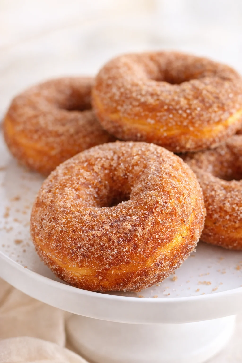 Golden donuts coated in cinnamon sugar on a white cake stand, close-up.