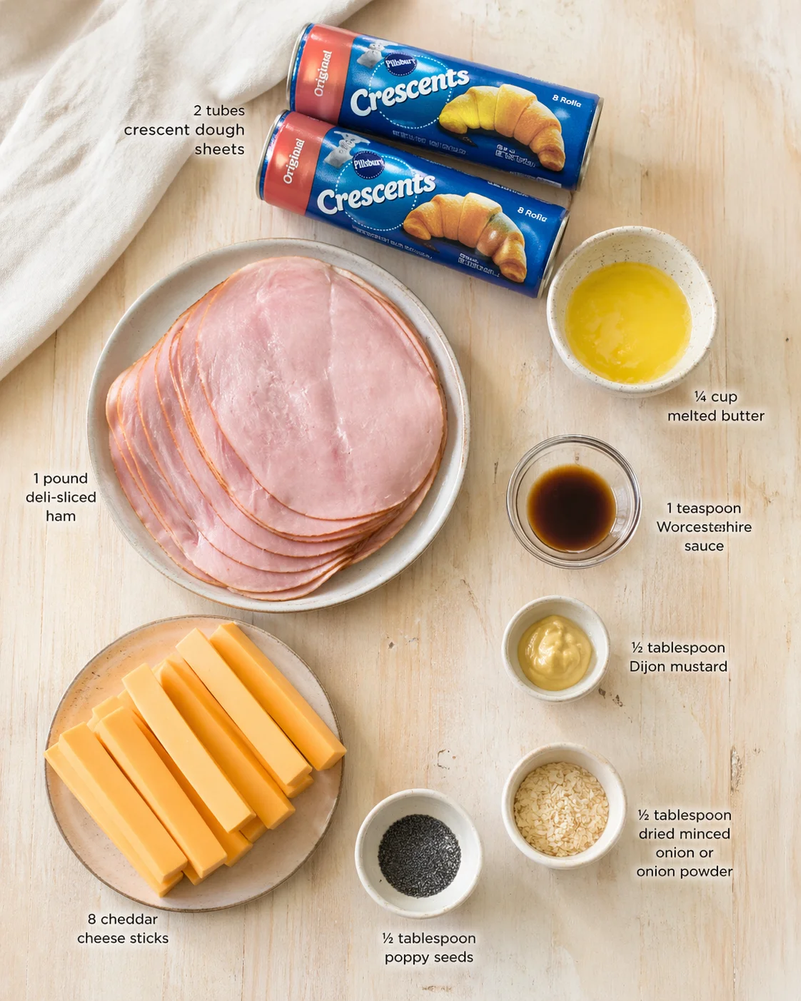 Overhead view of ingredients for a ham and cheddar crescent bake: crescent dough, ham, cheddar sticks, melted butter, Worcestershire sauce, Dijon mustard, poppy seeds, and onion powder.