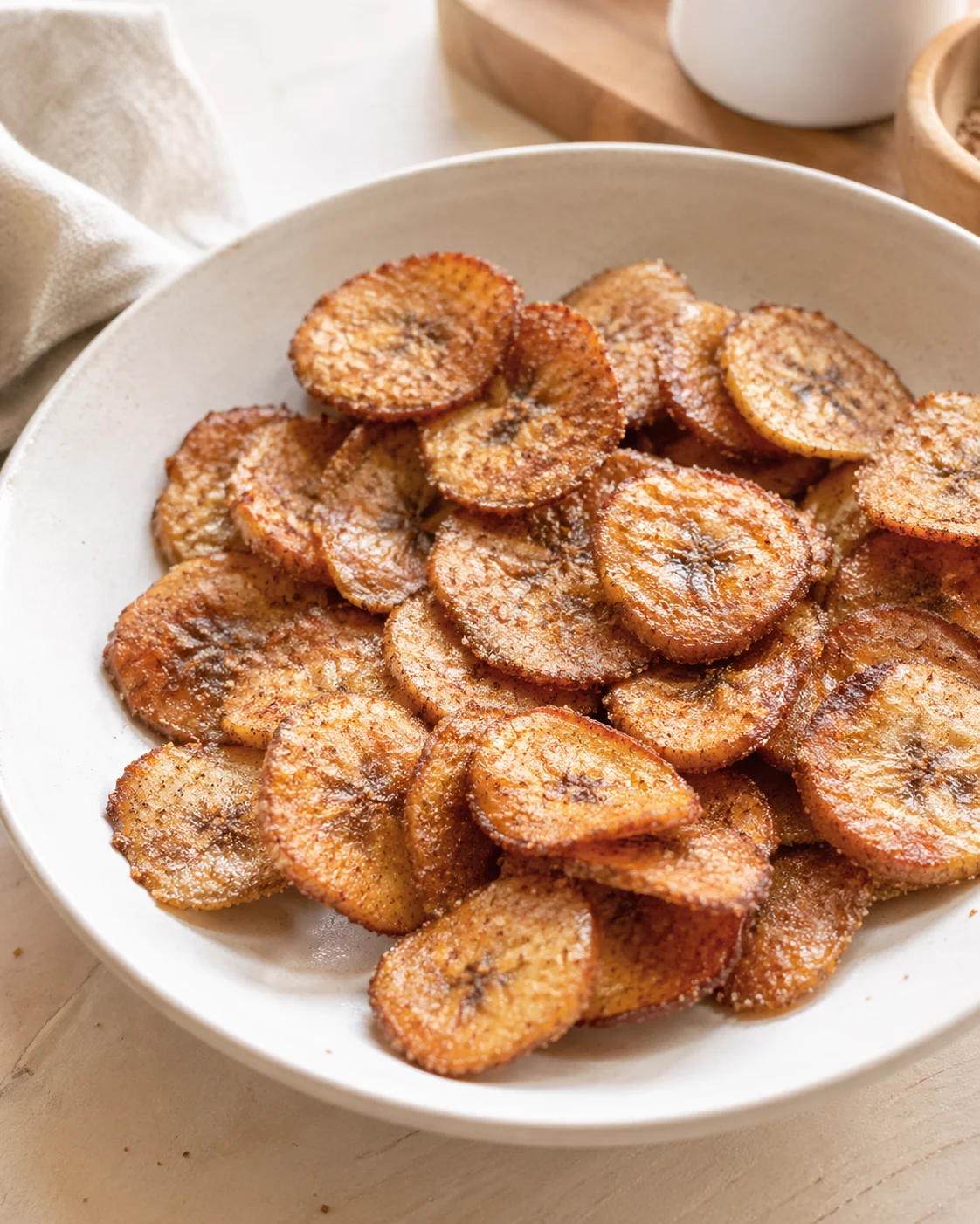 Golden-brown fried banana chips piled in a white bowl on a light wooden surface.