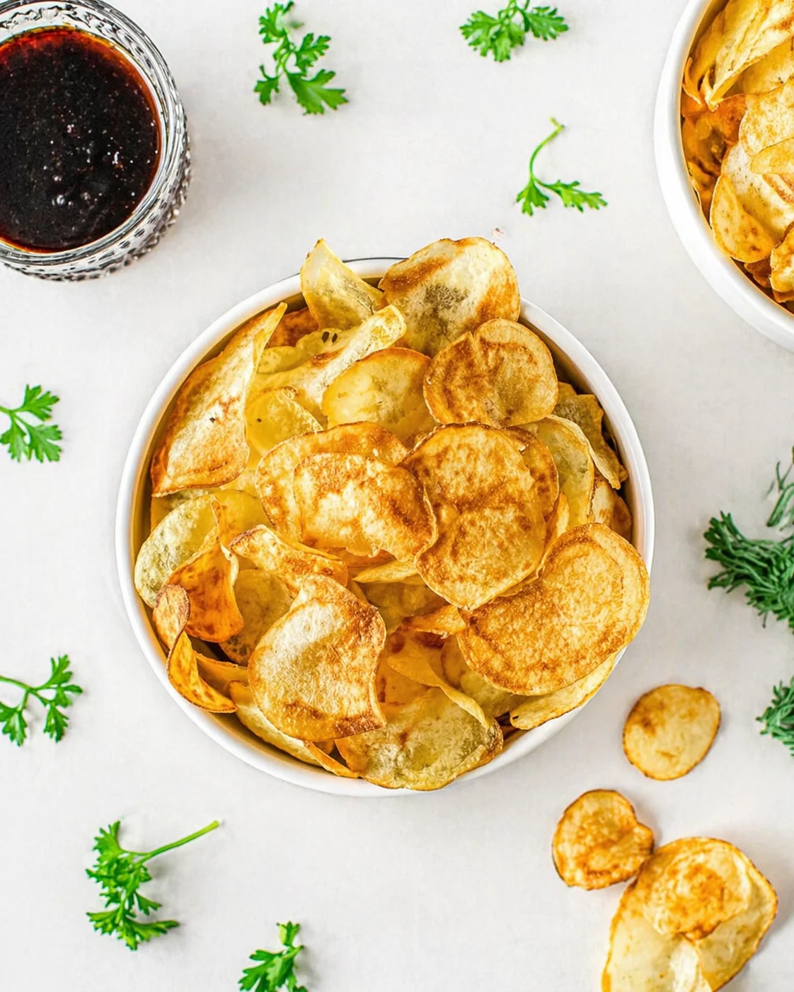 Round white bowl of golden potato chips on a light surface, with parsley and dip.