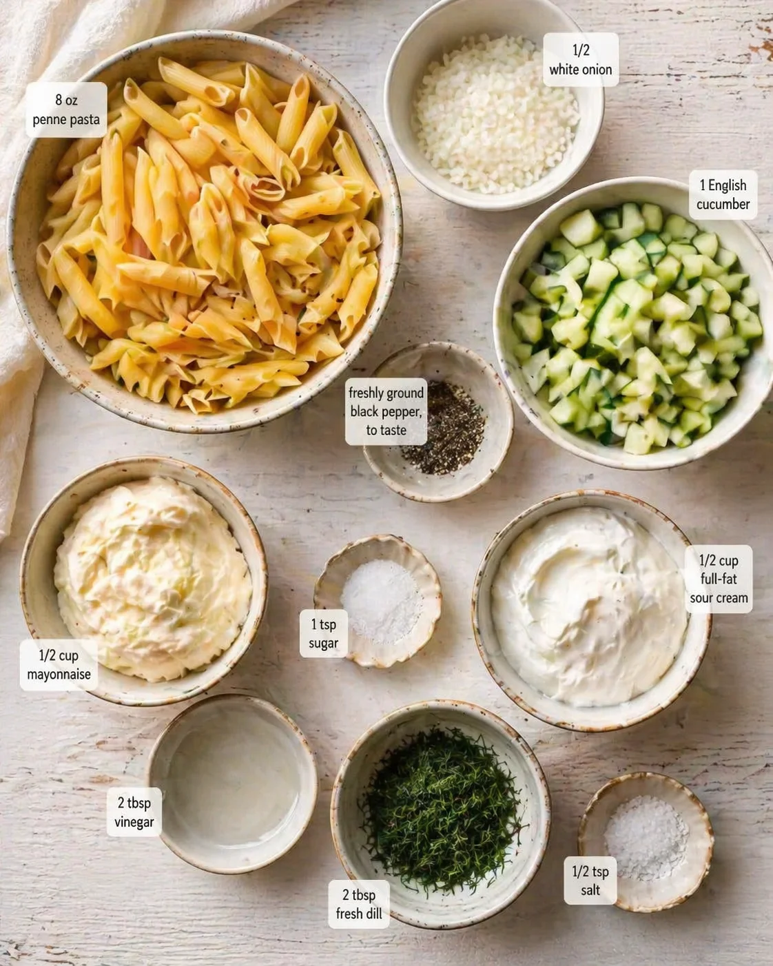 Top-down view of pasta salad ingredients in bowls with labeled ingredients: penne, cucumber, onion, mayo, sour cream, dill.