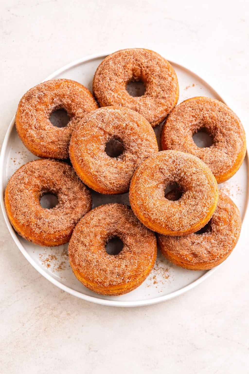 White plate holds a circle of cinnamon-sugar donuts, arranged neatly on a light background.