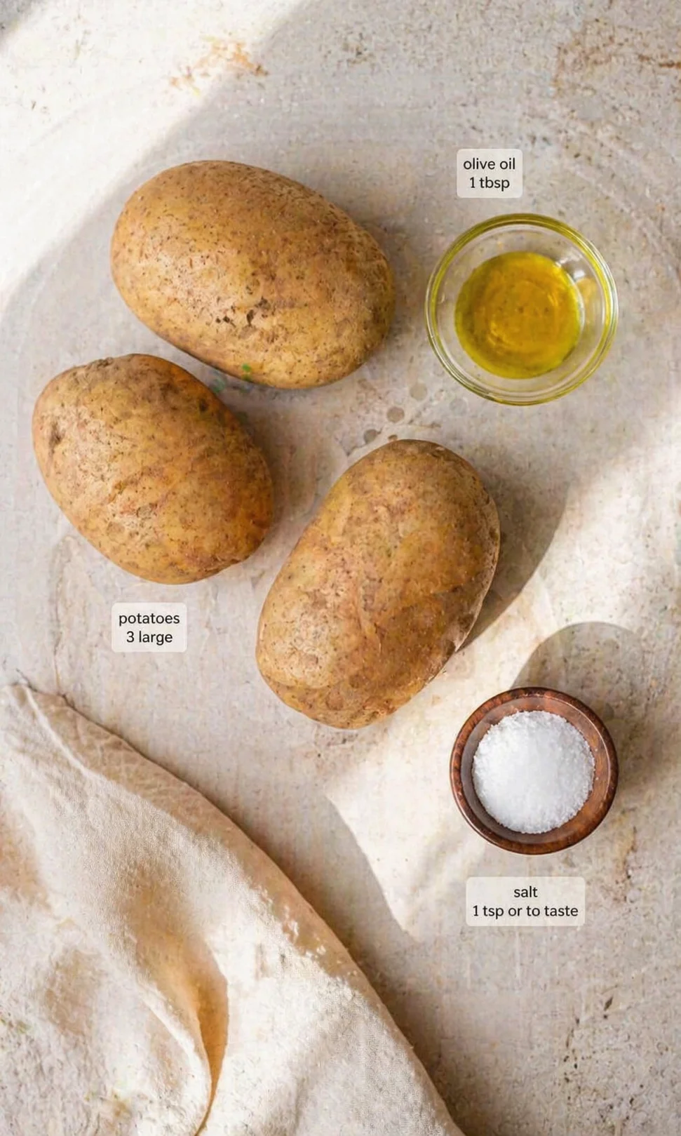 Three large potatoes with olive oil and salt on a textured neutral surface.