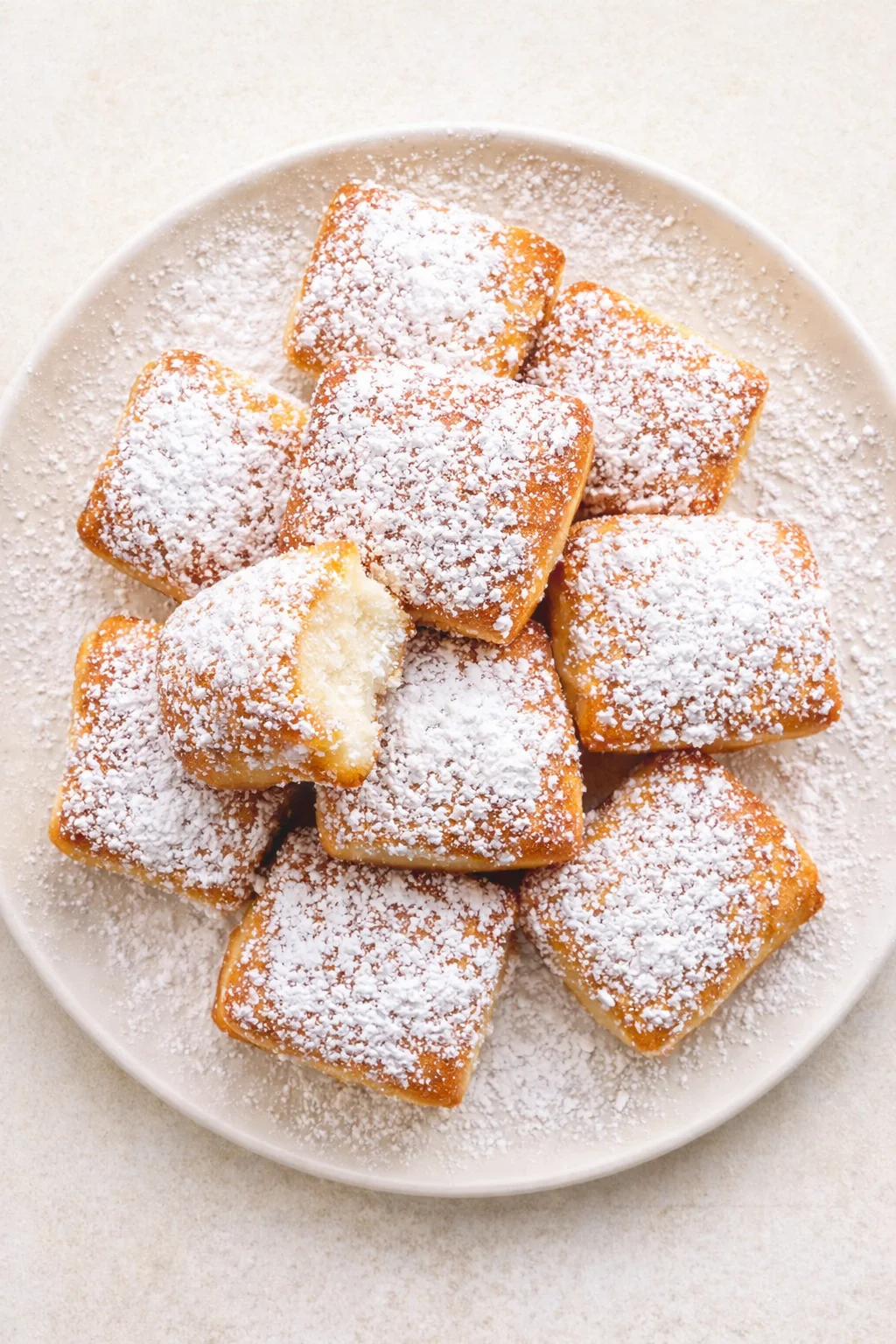 Pile of square, golden fried pastries dusted with powdered sugar on a white plate.