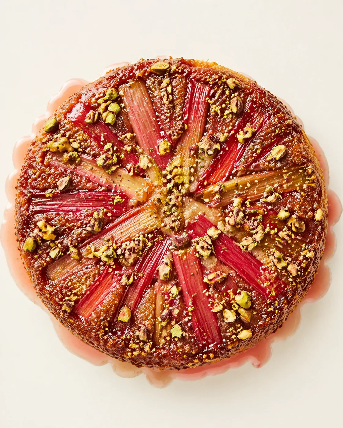 round dessert cake with rhubarb slices in a radial pattern, pistachio crumble, and pink glaze on a pale background