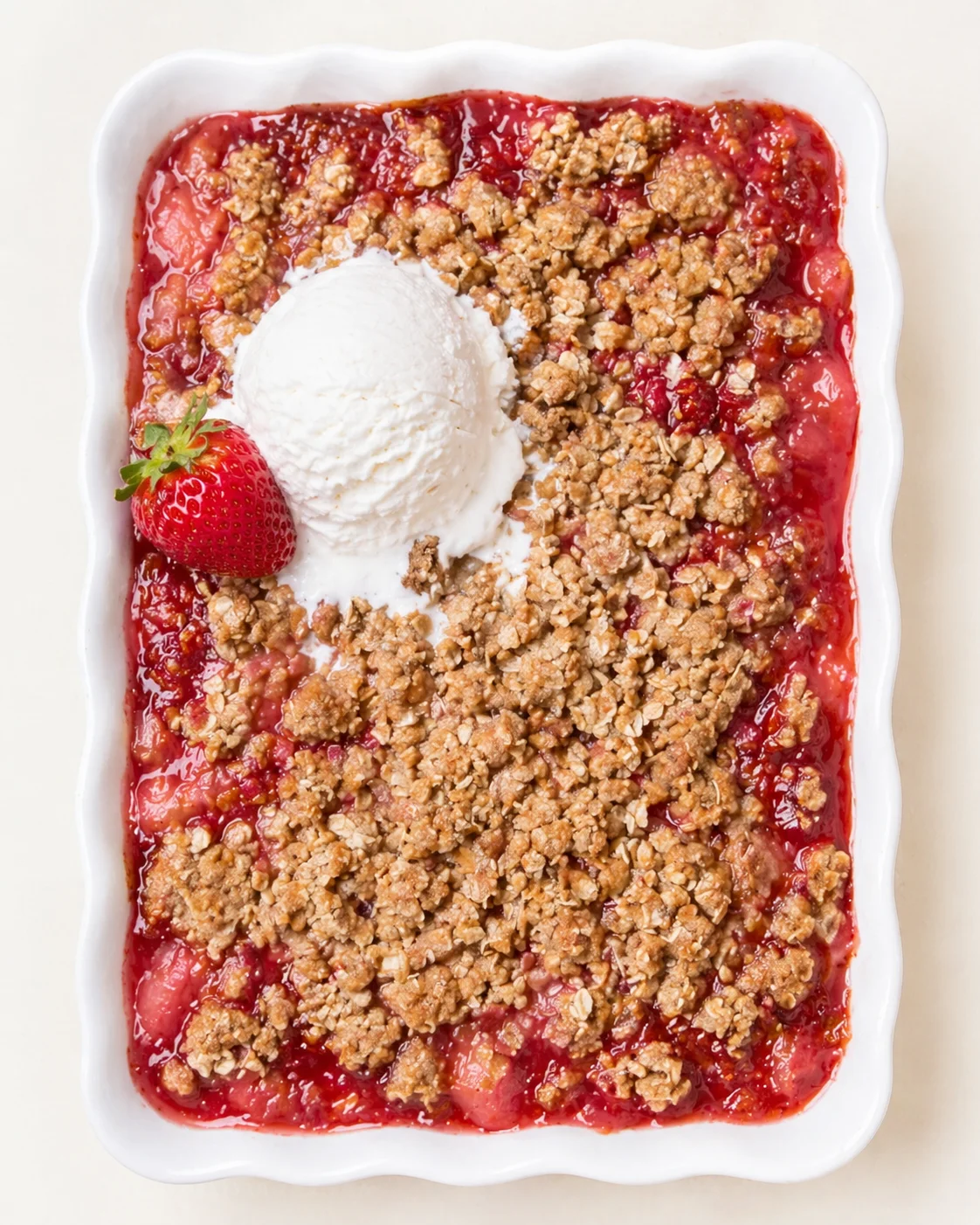White scalloped baking dish of strawberry crumble topped with vanilla ice cream and a strawberry.